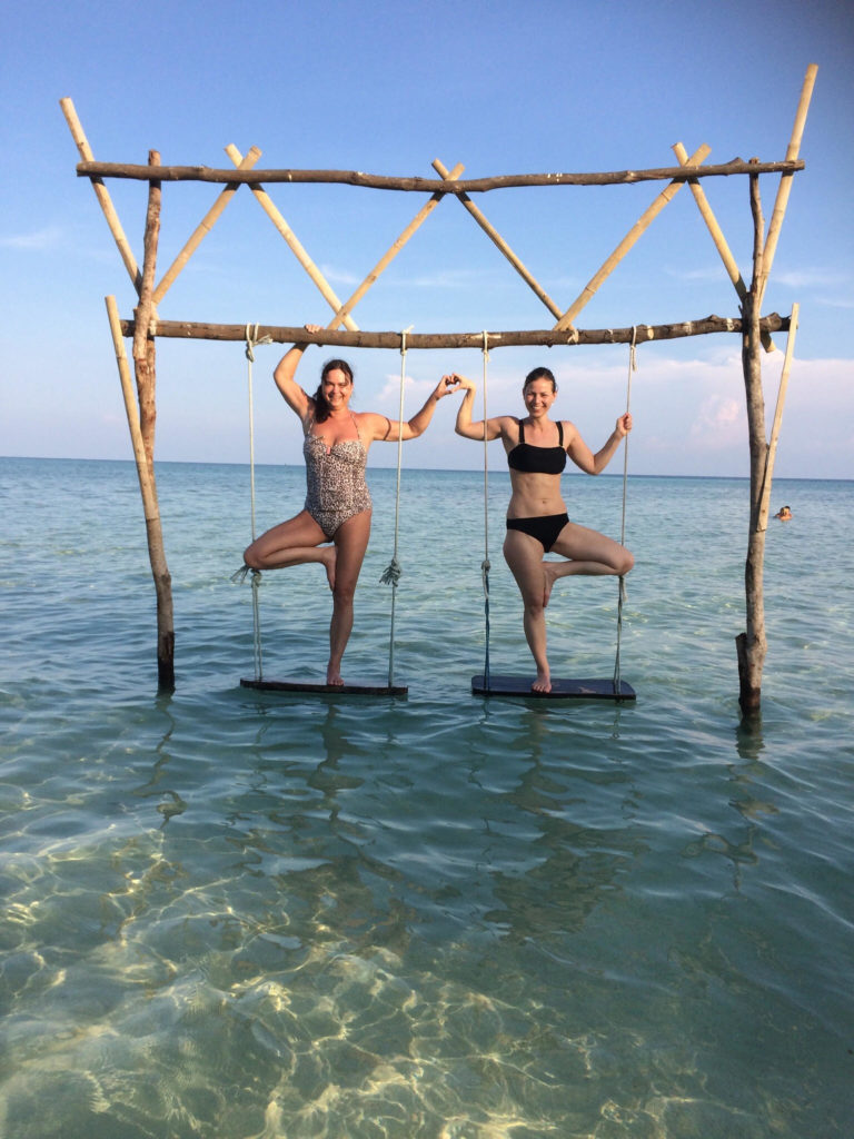 Two women stand on swings above crystal blue water, mirroring each others' pose